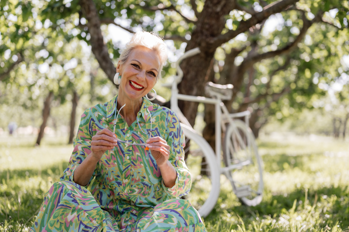 Older woman wearing a dress and sitting in a field smiling, with a bike behind her leaning against a tree